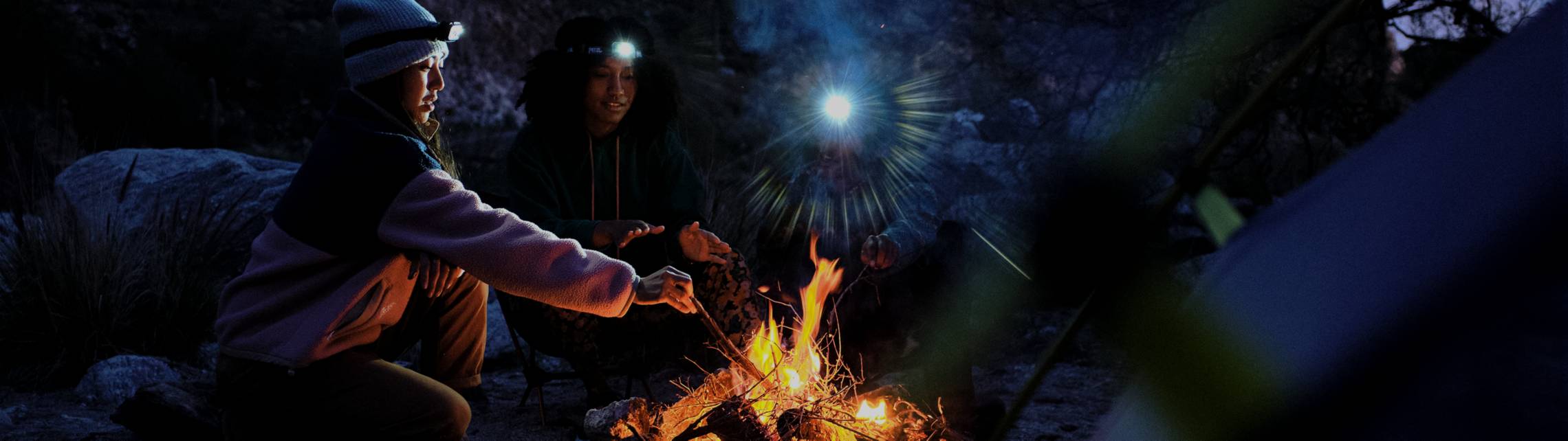 Two women wearing headlamps dispersing light tend to a campfire at night.