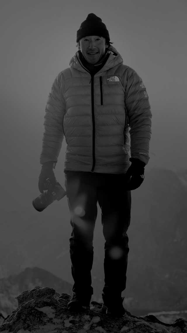 Athlete and filmmaker, Jimmy Chin, stands on a summit with camera in hand.