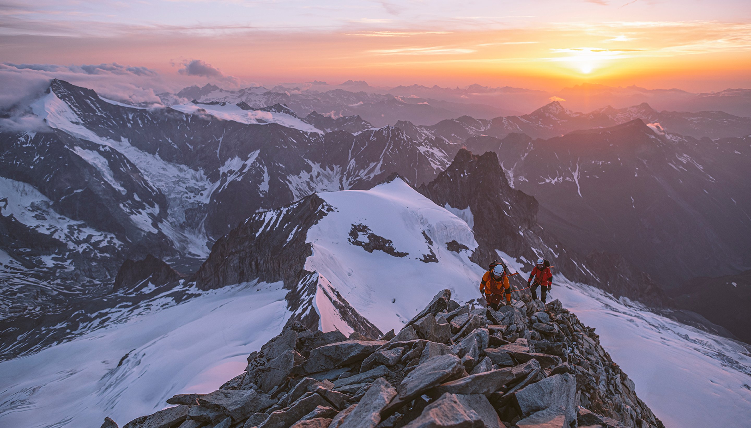 In the remote alpine, athletes scale a snowy peak at magic hour.