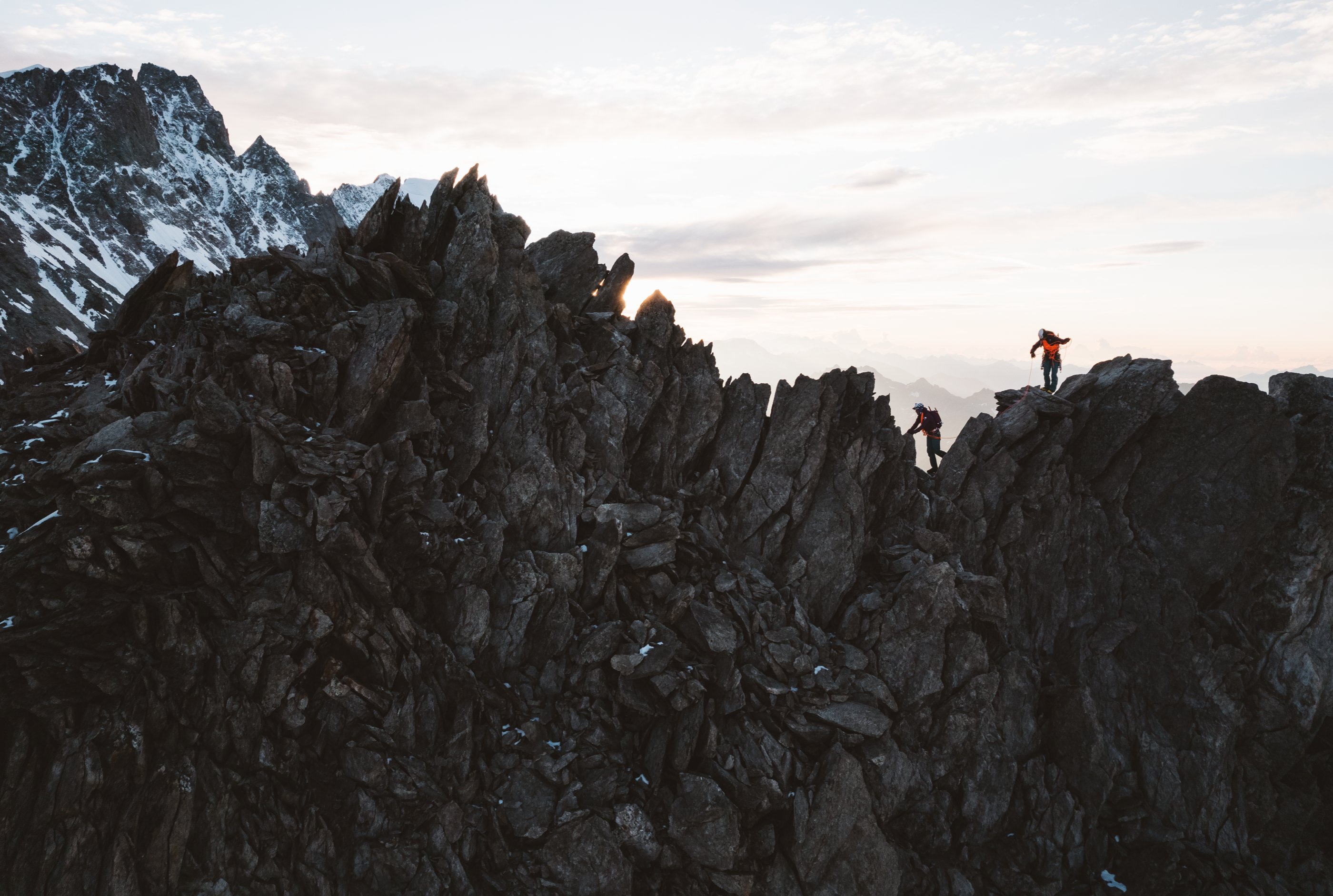 Two intrepid mountaineers are silhouetted against the cold, gray sky as they scramble across a jagged alpine ridge.