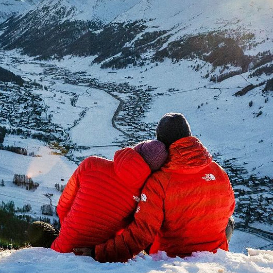 A couplewearing red puffy jackets from The North Face sit on a snowy summit looking down on a valley.