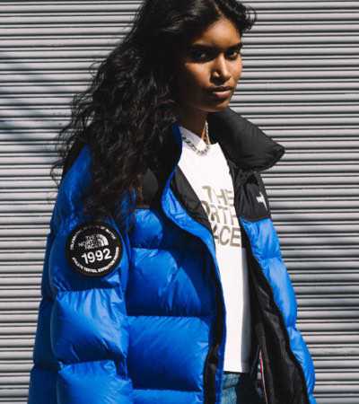Standing against a wall wearing the new blue ’92 Nuptse, a woman holds the Nuptse anniversary badge up to the camera.