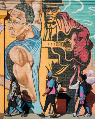 Trois athlètes de l’équipe portant des vêtements de la collection Black History Month de The North Face marchent
            devant une peinture murale.