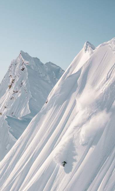 A distant image of a snowboarder descending the steep face of snowy mountains.
